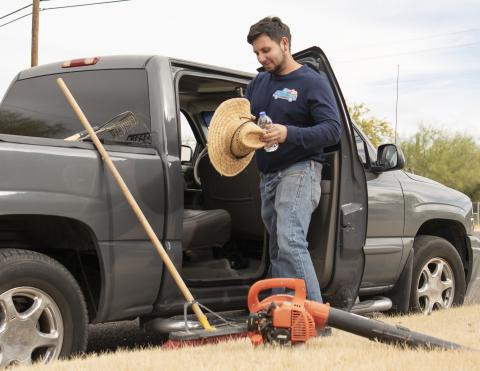 Un hombre con una camisa azul de manga larga y jeans, sosteniendo un sombrero de sombra y parado junto a una camióneta gris, con herramientas de jardinería sentadas en el suelo.