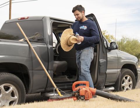 Un hombre con una camisa azul de manga larga y jeans, sosteniendo un sombrero de sombra y parado junto a una camióneta  gris, con herramientas de jardinería sentadas en el suelo.